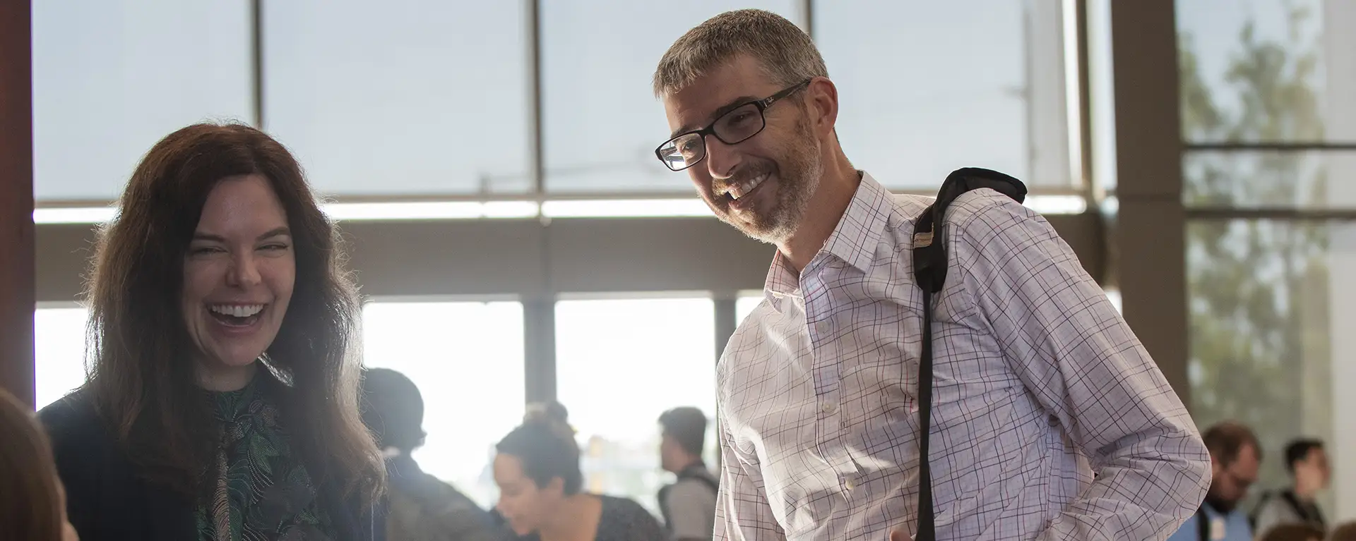 A student carrying a laptop bag smiles while chatting with another student.