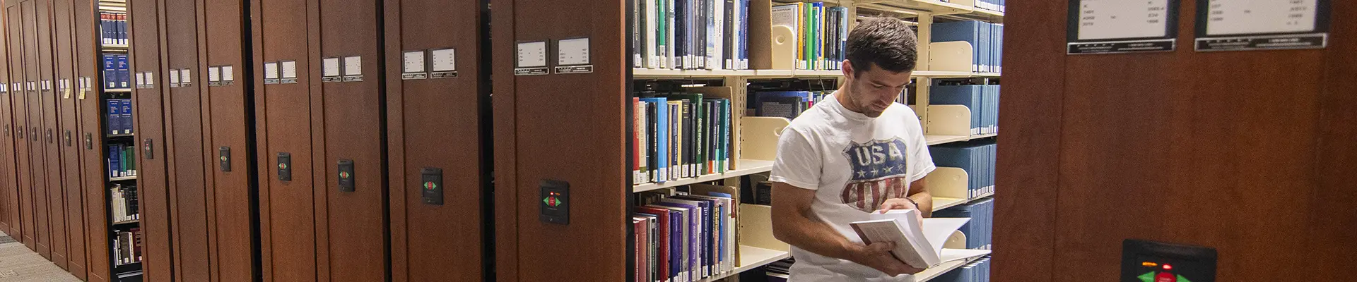 A student flipping through a book in the library stacks.