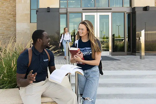 two students seated at table in lobby smiling while looking at laptop computer.