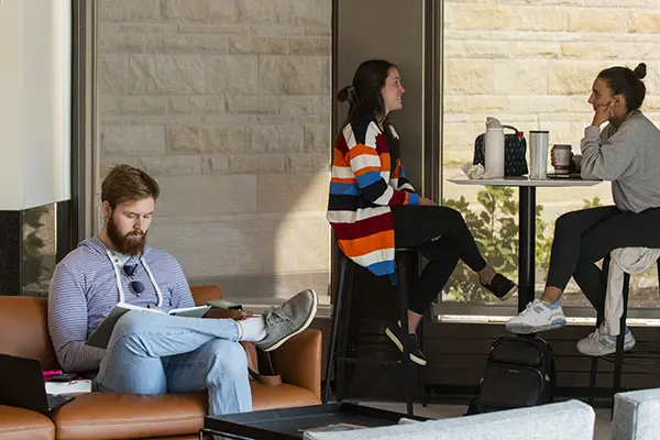 Two students talk at a table while another studies on a couch in the lobby.