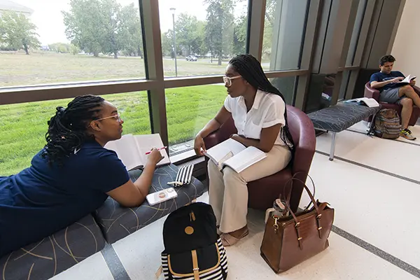 two students sitting near window talking with books and papers