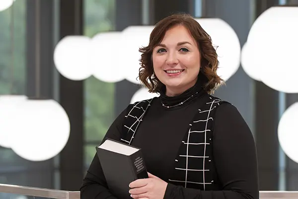 student standing and smiling holding a book in SOL building lobby balcony with pendant lights hanging behind.