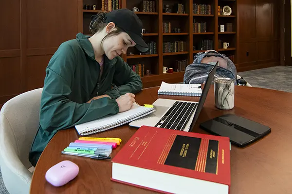 A student takes notes while studying with a laptop and book on the table before her.