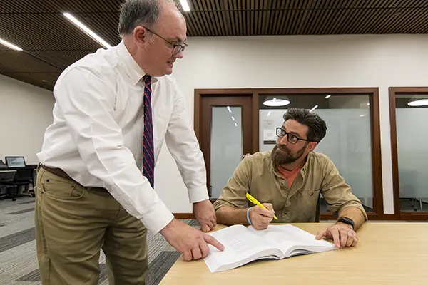 A professor helps a student taking notes out of a book in the library.