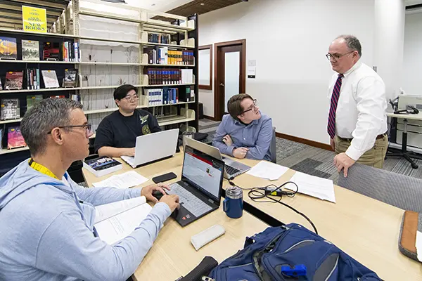 A professor talks with a group of students studying together in the library.